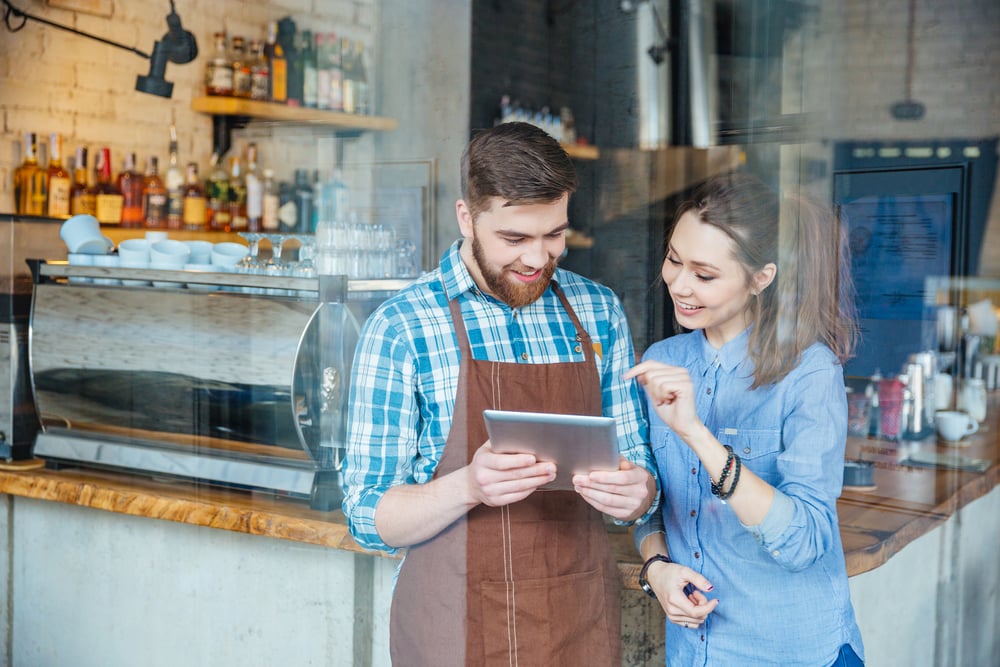 コーヒーショップでタブレットを持つ笑顔のハンサムなウェイターとそれを指差す若い美女