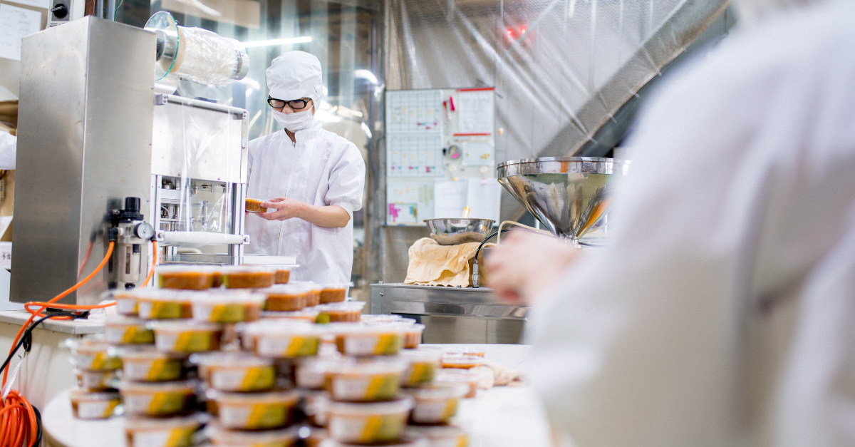 白衣にマスク姿の人物が、山積みにされた食品パッケージの後ろで蓋をしている。